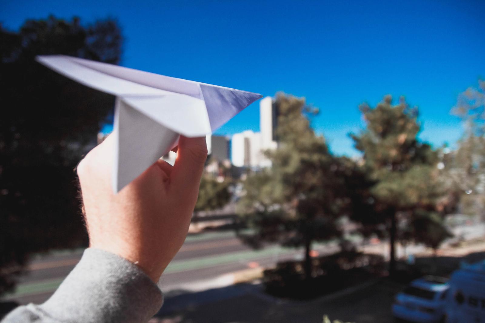 Una mano a punto de lanzar un avión de papel bajo un cielo azul con árboles y edificios de fondo