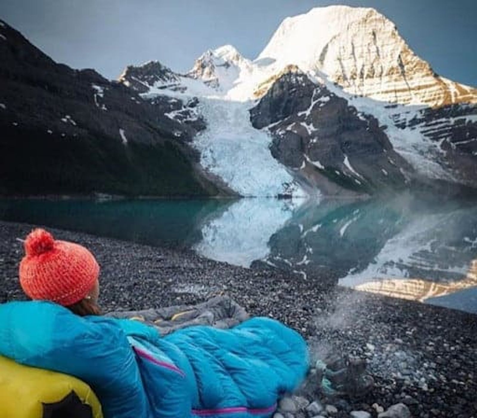 Persona con gorro y chaqueta azul recostada junto a un lago, frente a una montaña nevada y un glaciar.