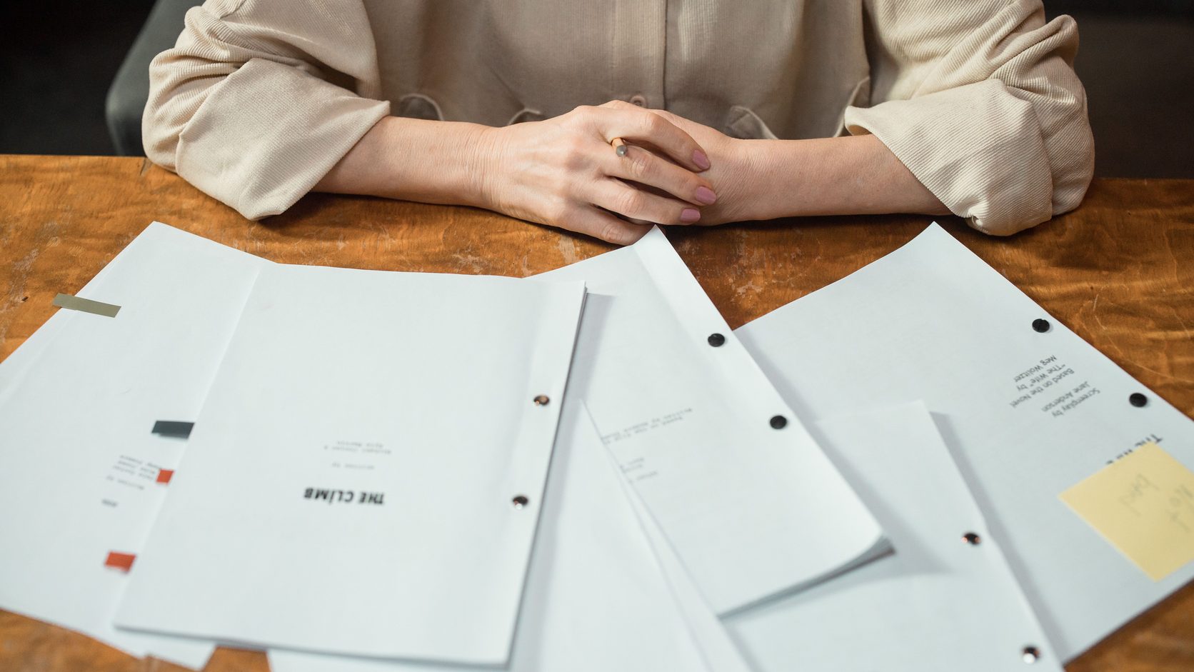 Printed documents spread across a wooden table, with a person's folded hands behind them
