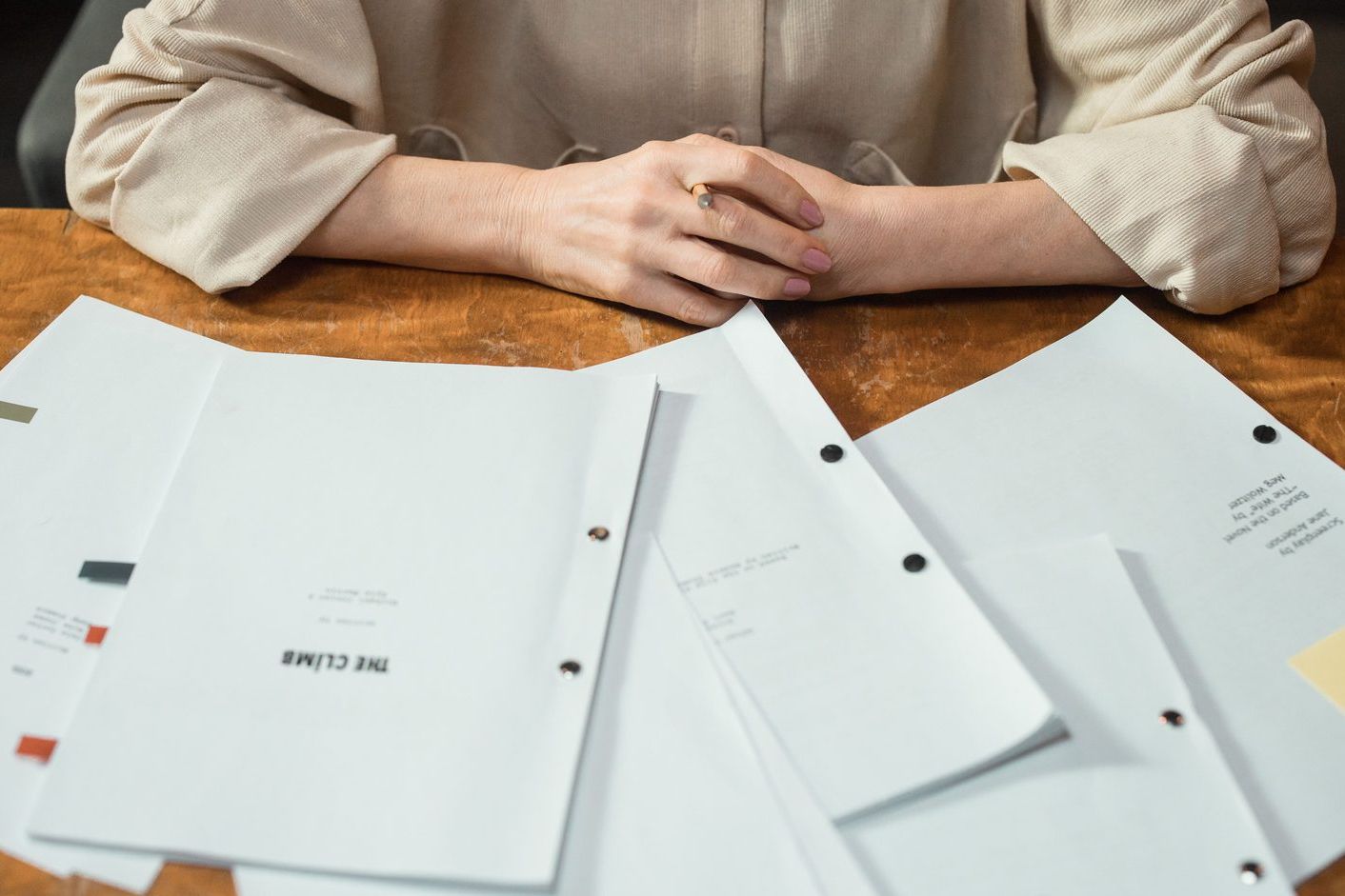 Printed documents spread across a wooden table, with a person's folded hands behind them