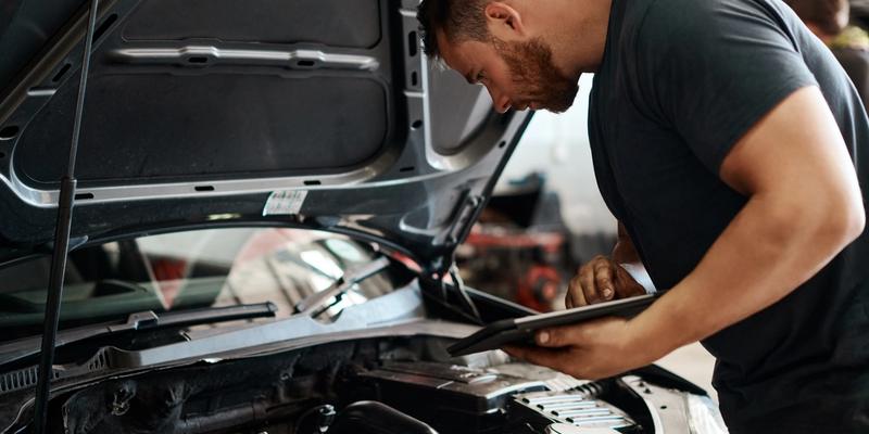 En mann i grå t-skjorte med en ipad i hånda som står bøyd over en bil med panseret åpent og undersøker bilens motor.
