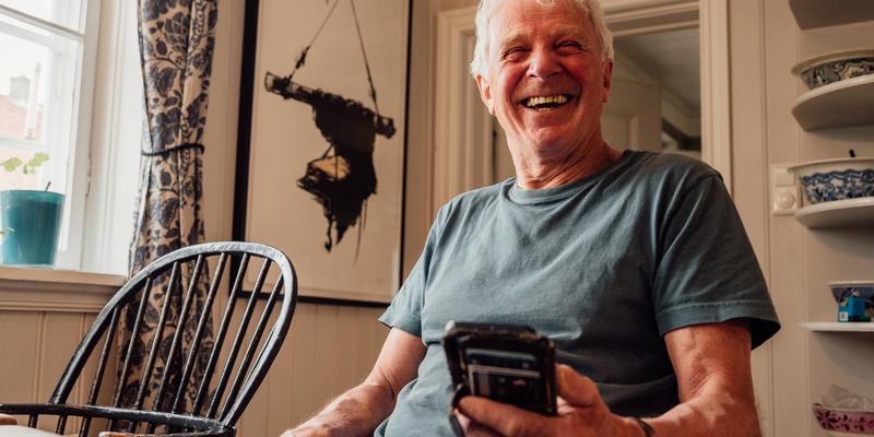 En mann som sitter ved et kjøkkenbord og holder telefonen i hånda mens han smiler mot kamera