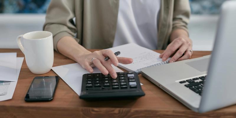 Person with a white t-shirt and beige shirt, sitting at a desk in front of a computer with papers, mobile phone, coffee cup who is using a calculator.