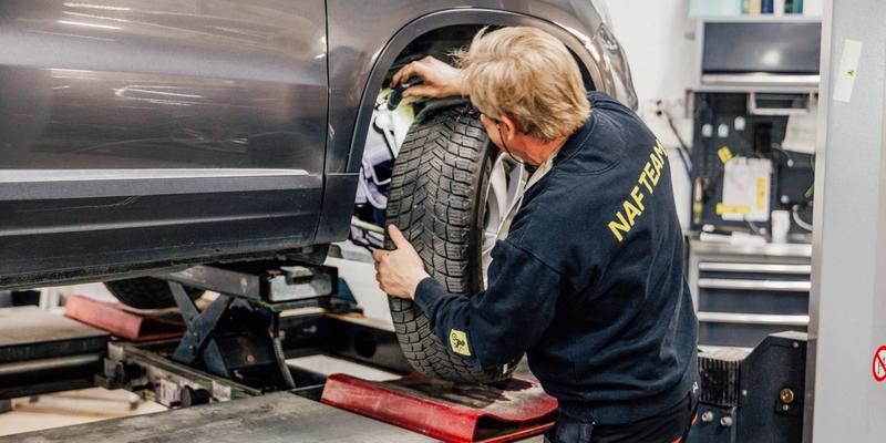 En mann i svart genser og arbeidsbukse på et verksted som inspiserer en bil og bak hjulet for å sjekke bilens tilstand.
