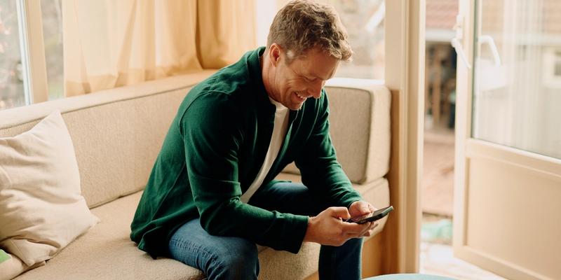 En mann i grønn skjorte som sitter på en beige sofa og ser på mobilen sin mens han smiler.