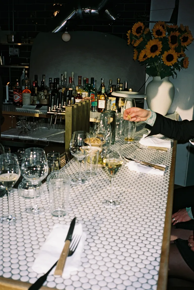 table setting on Asters bar with a hand reaching out. Bottles and a pot with sunflowers in the background