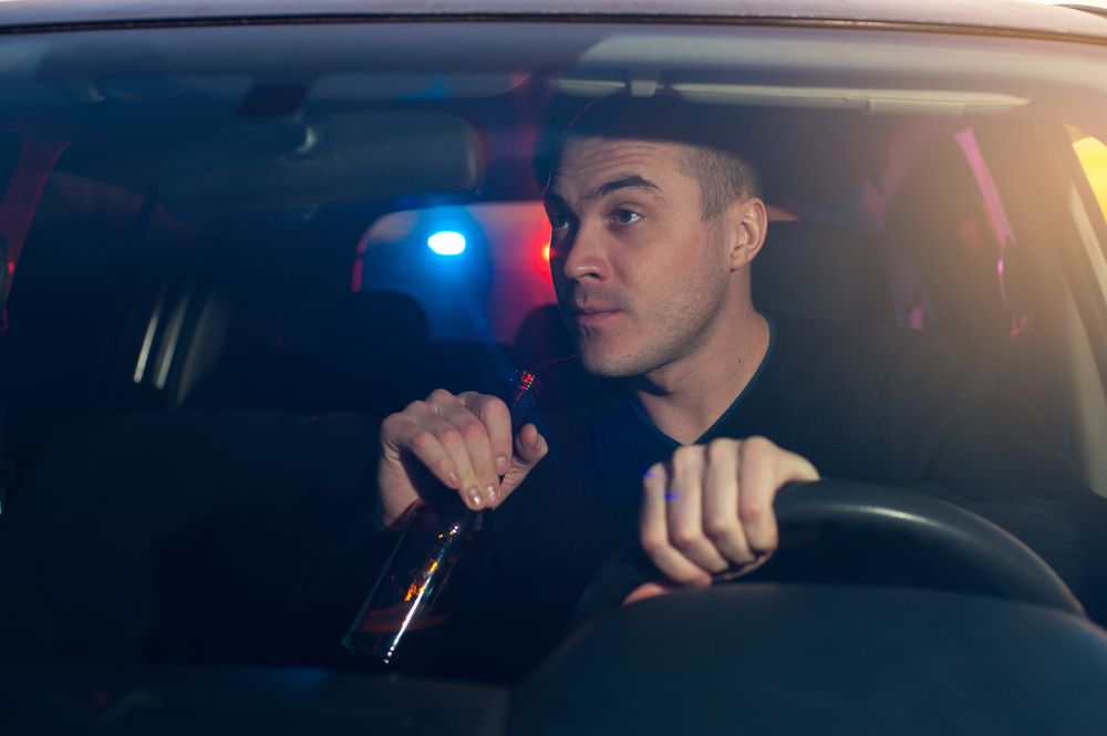 Driver holding a beer bottle behind the wheel during a nighttime DUI traffic stop with police lights in the background
