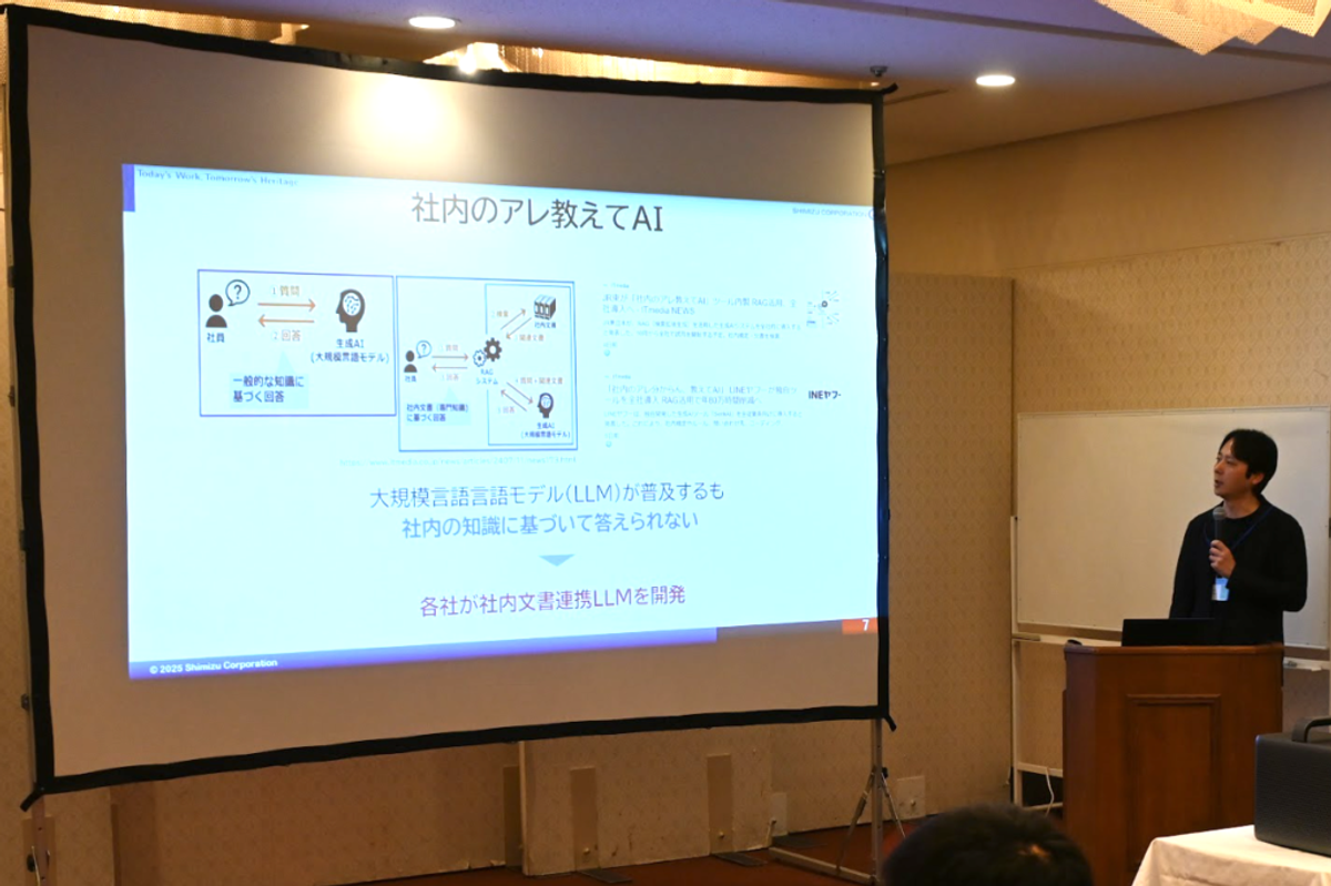 清水建設による発表スライドで、「社内のアレ教えてAI」というタイトルの下に、社内知識を活用したAIシステムの概念図が表示されている。スライドには「大規模言語言語モデル(LLM)が普及するも社内の知識に基づいて答えられない」という課題と、「各社が社内文書連携LLMを開発」という解決策が示されている。講演者が壇上で説明している様子。