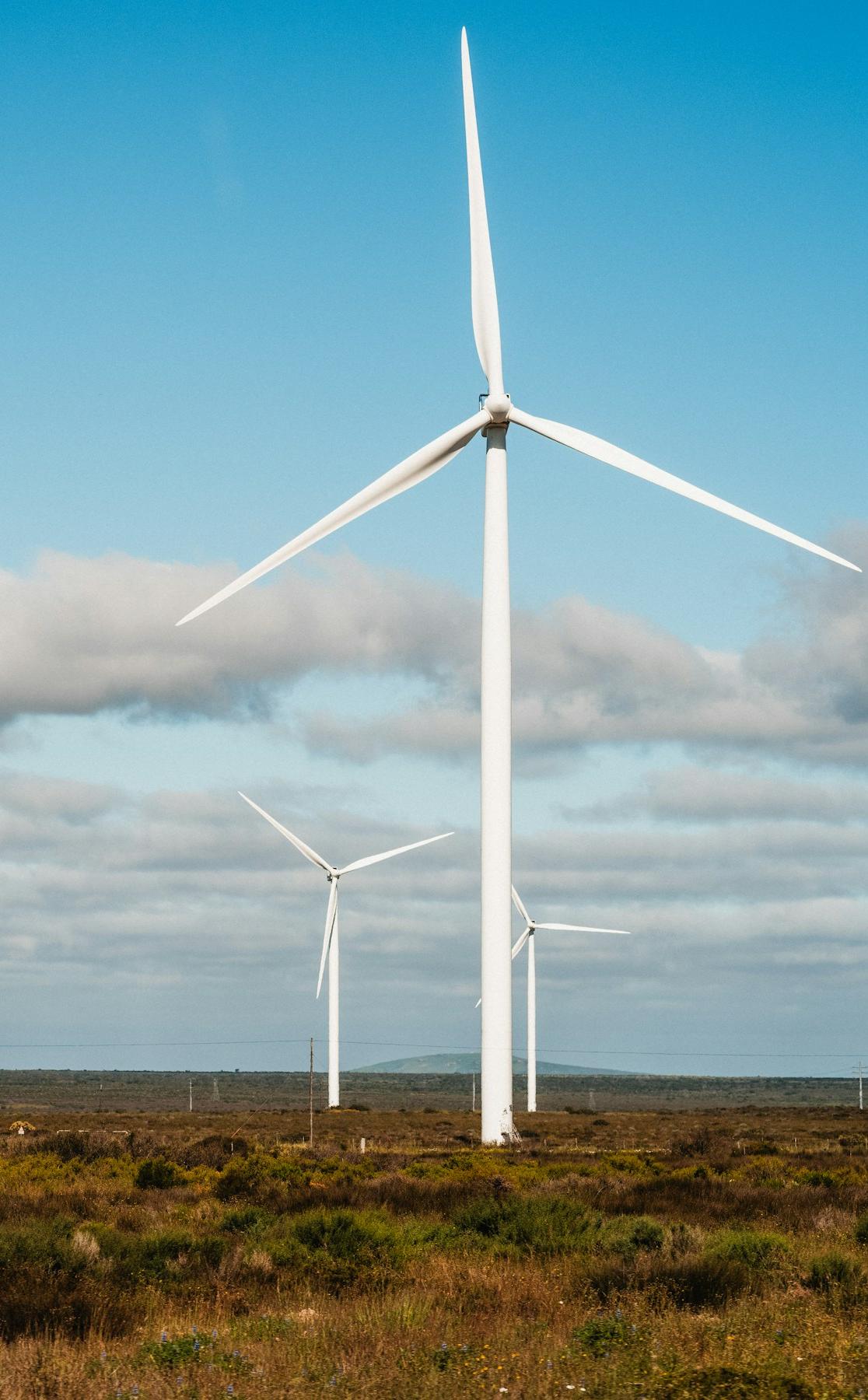 Wind turbines standing over dry scrubland under a cloudy blue sky.