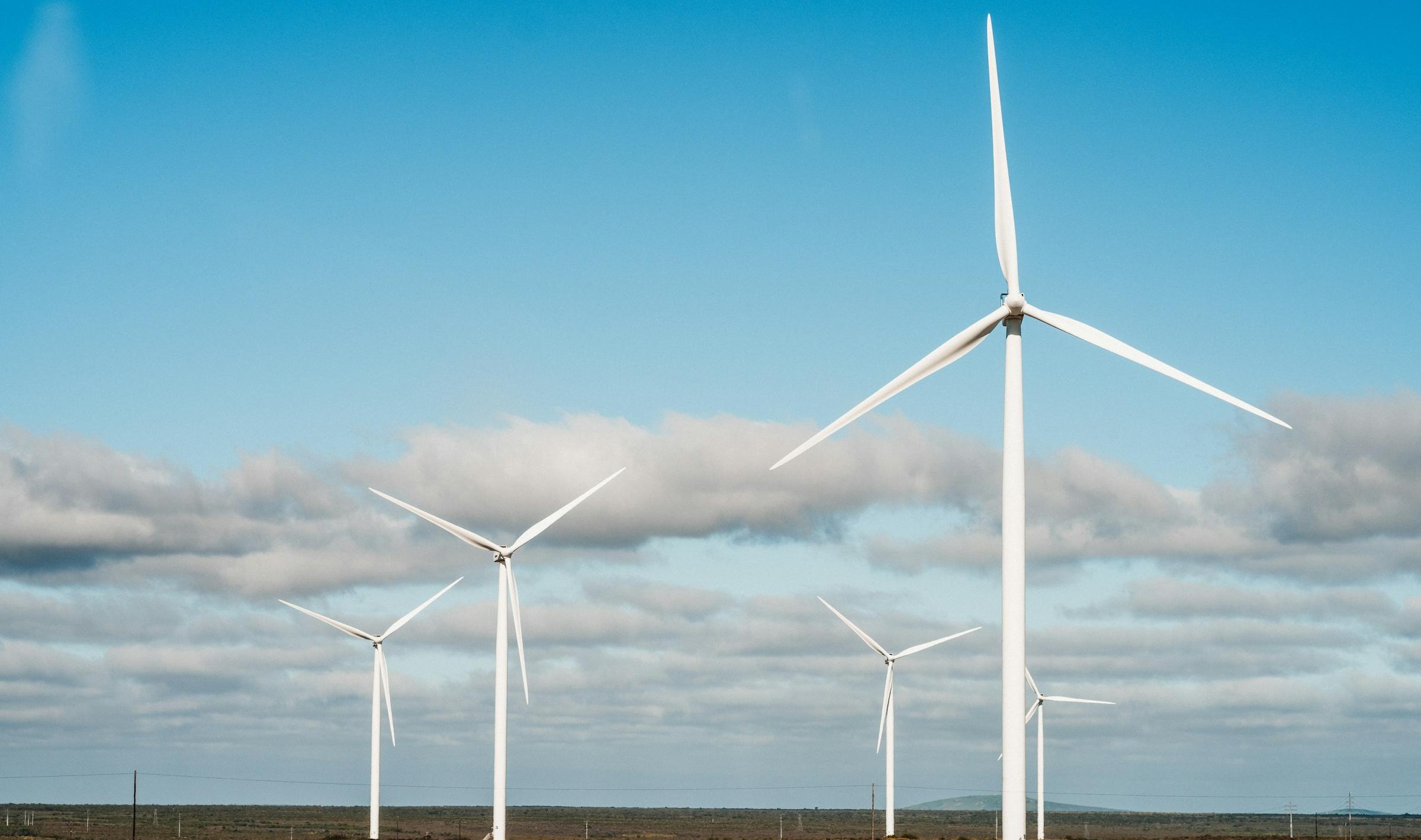 Wind turbines standing over dry scrubland under a cloudy blue sky.