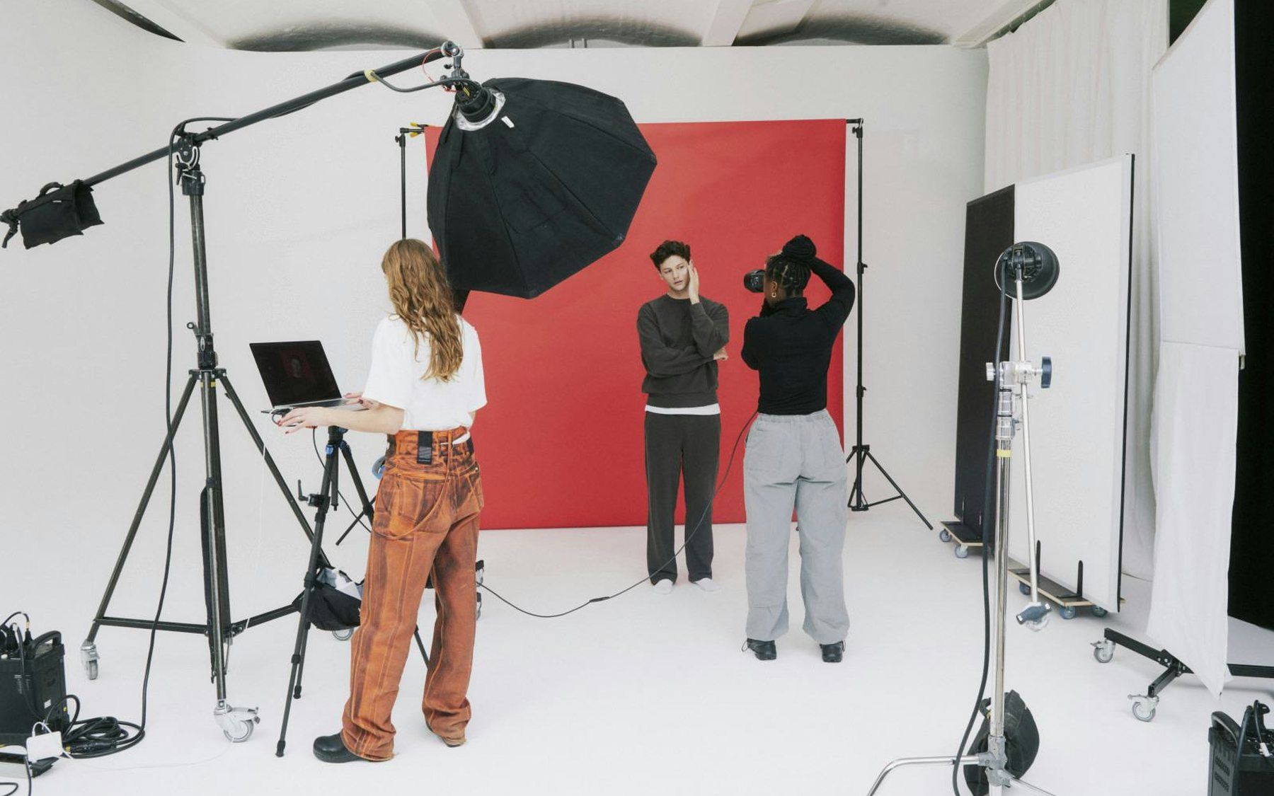 A photography studio in use: lighting stands, a softbox and a red paper backdrop, with a crew member at a laptop and two models waiting.