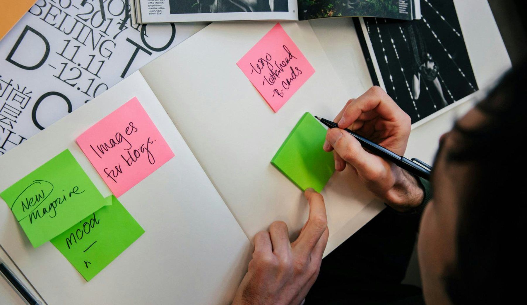 Hands arranging green and pink sticky notes across an open workbook, surrounded by printed type samples and photographs.