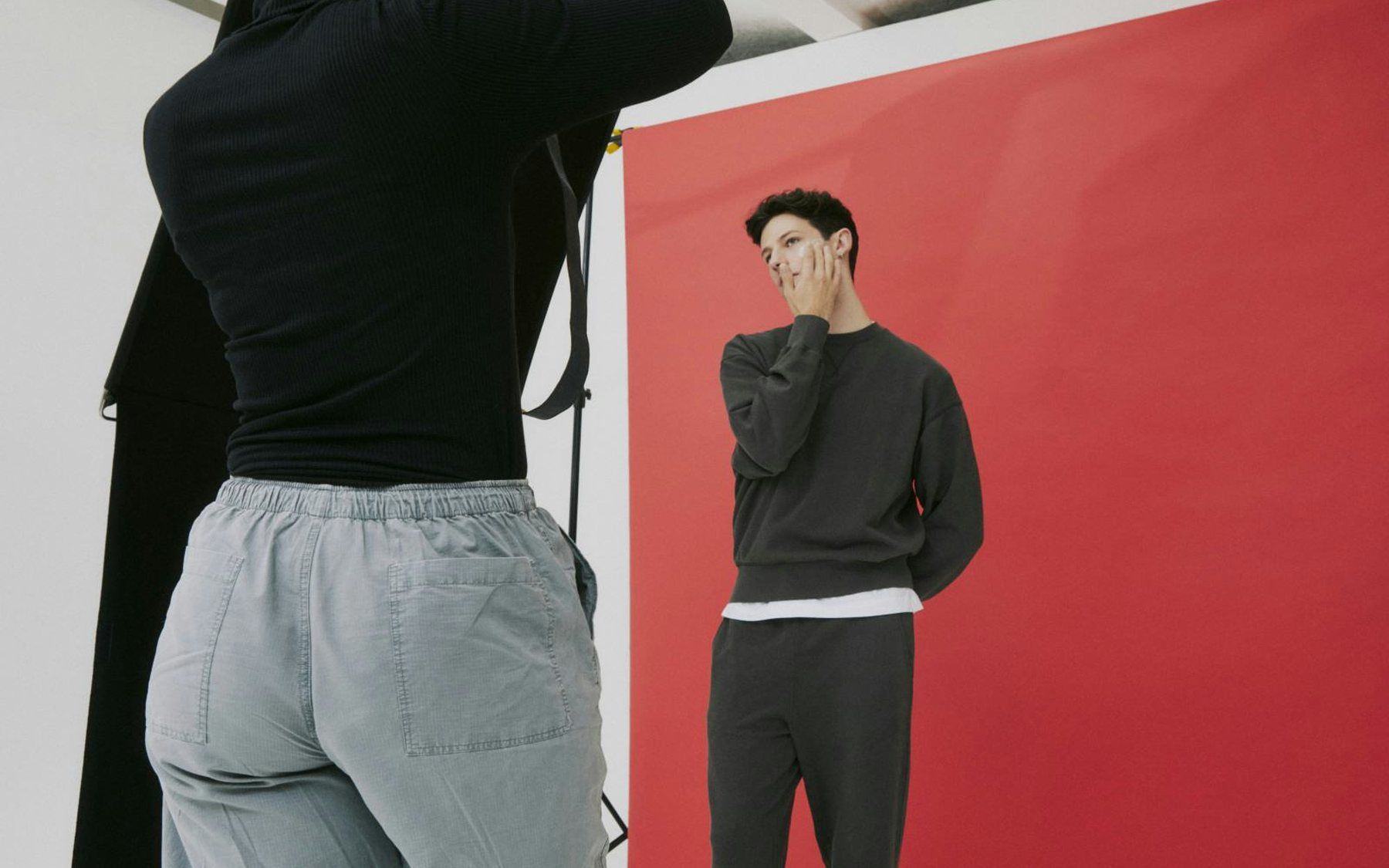 A model in grey and black standing against a red backdrop, seen past the shoulder of the photographer in the foreground.
