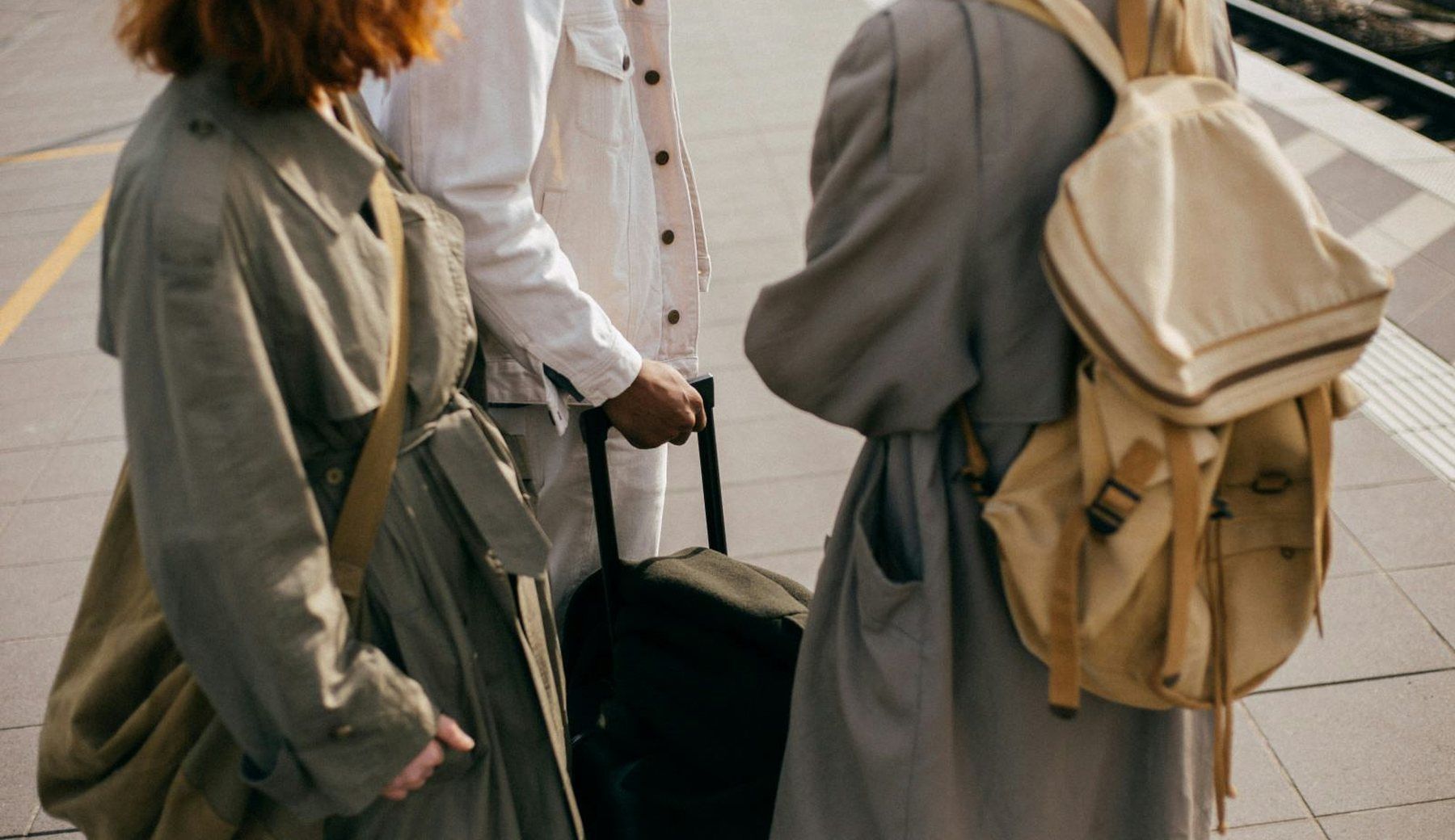 Three travellers photographed from behind on a station platform, carrying backpacks and wheeled luggage, in muted trench coats.