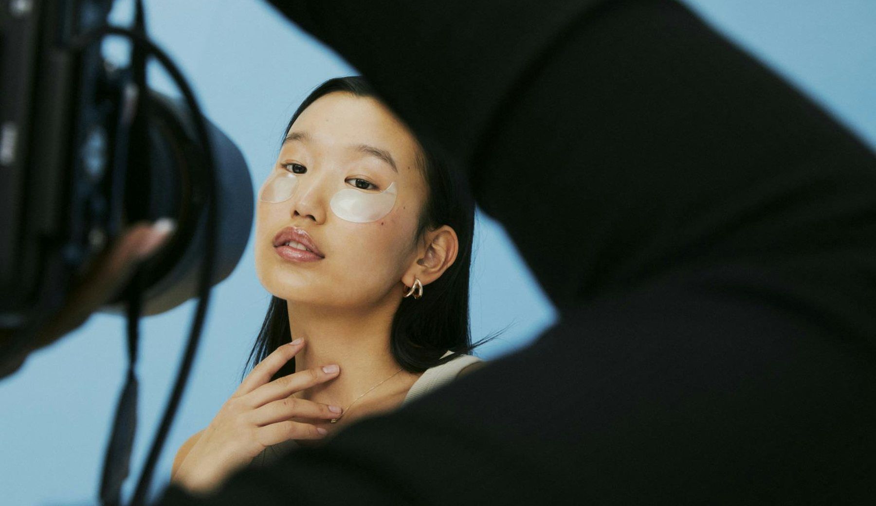 A portrait of a woman wearing under-eye patches, hand raised to her chin, against pale blue, with a camera rig visible at the edge of the frame.