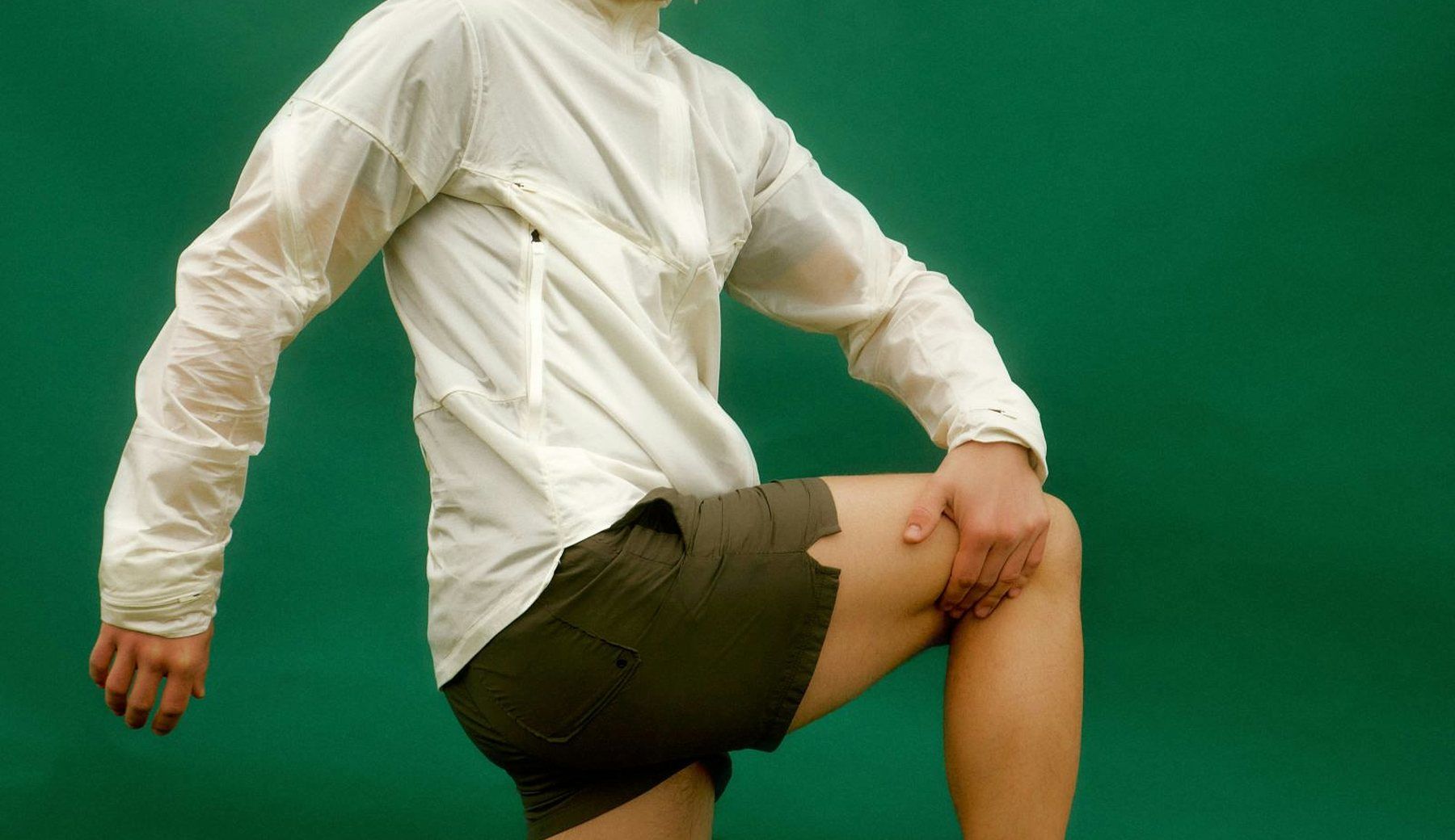 A figure in a white jacket and shorts, one leg raised, against a flat green studio backdrop.