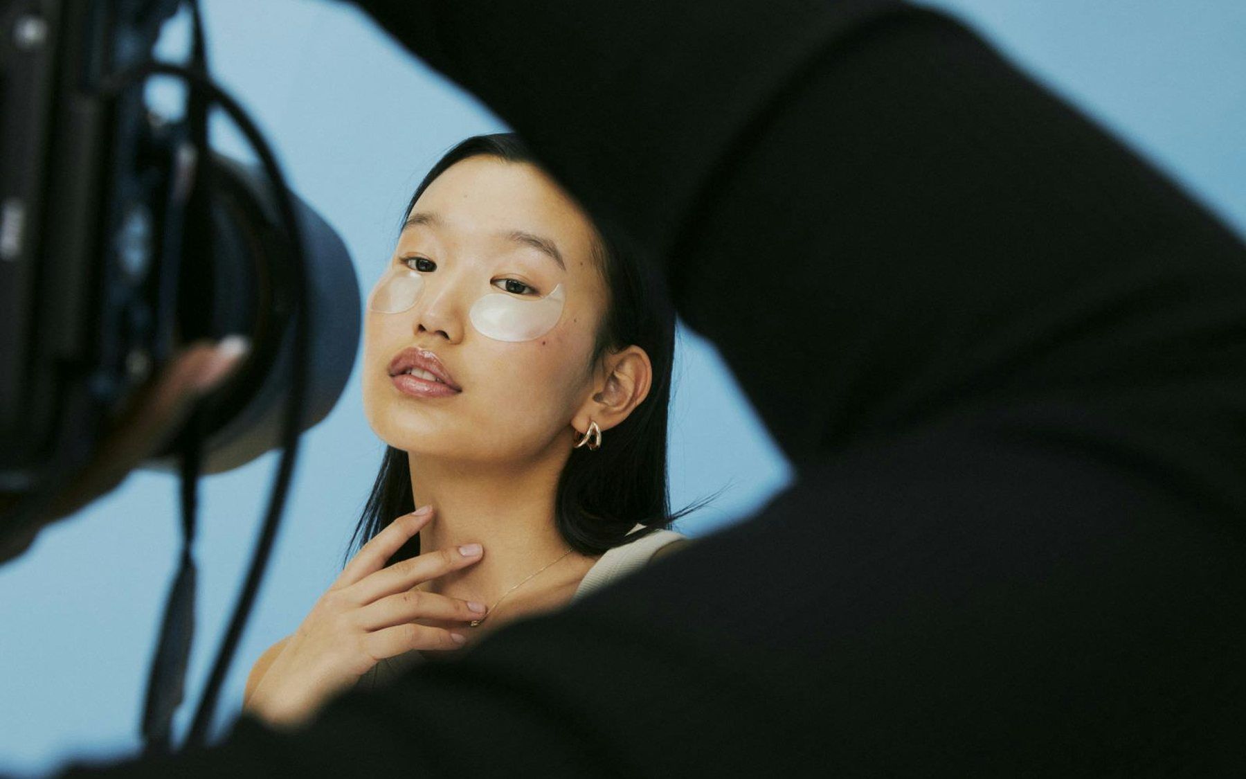 A portrait of a woman wearing under-eye patches, hand raised to her chin, against pale blue, with a camera rig visible at the edge of the frame.