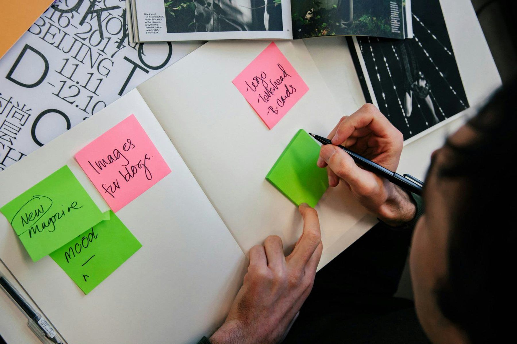 Hands arranging green and pink sticky notes across an open workbook, surrounded by printed type samples and photographs.
