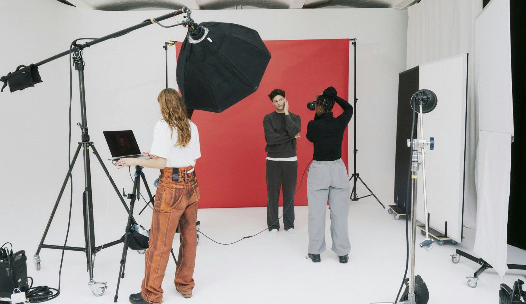 A photography studio in use: lighting stands, a softbox and a red paper backdrop, with a crew member at a laptop and two models waiting.