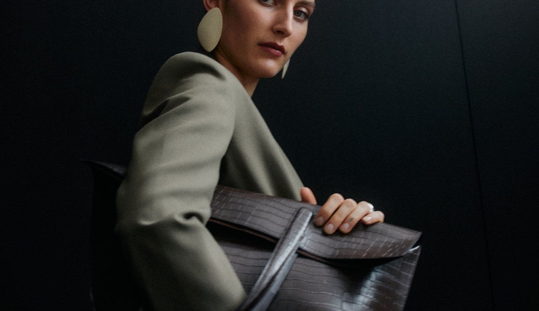 A model in an olive jacket and a large disc earring, turning to look back over her shoulder, holding a dark textured bag against a black ground.