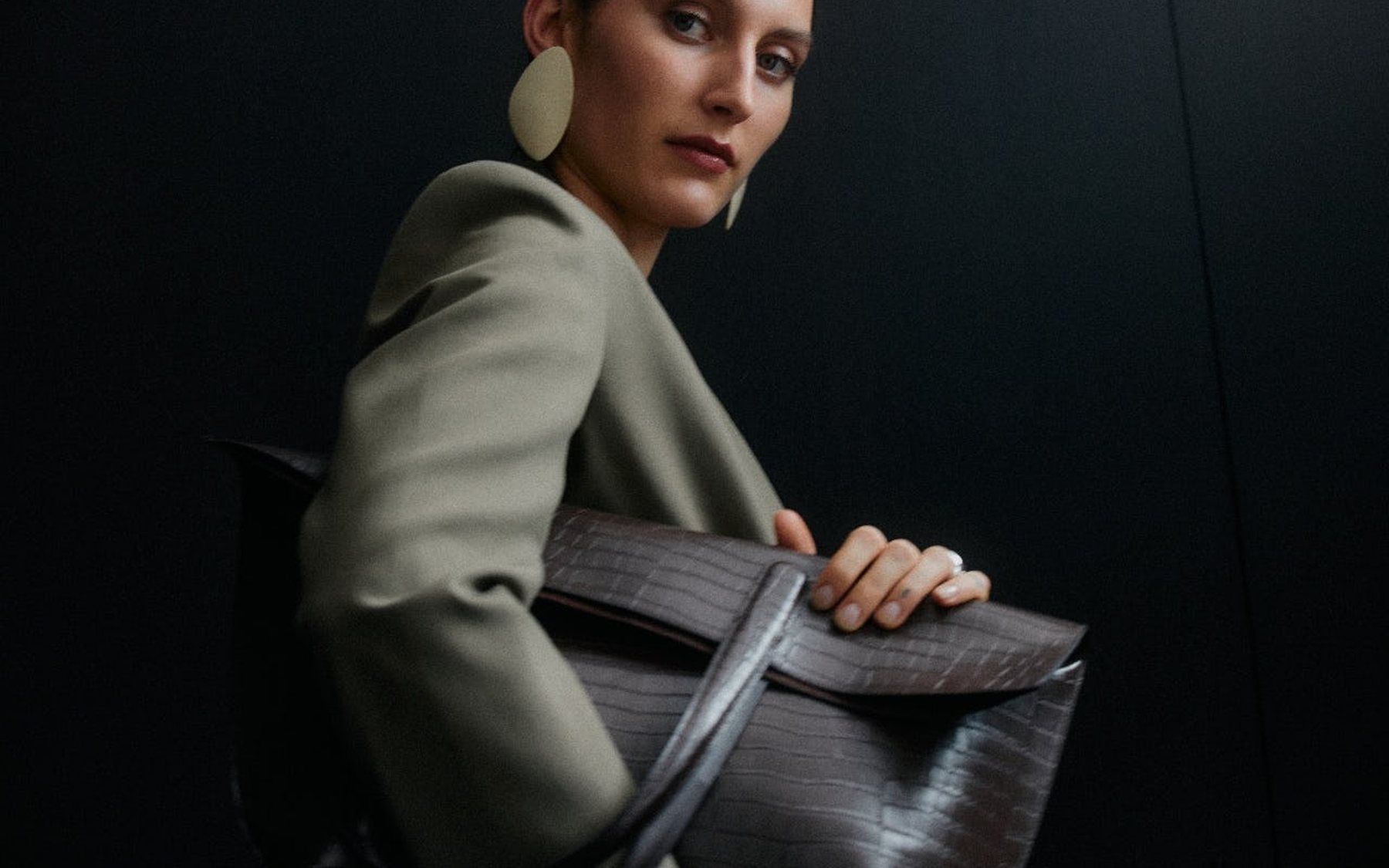 A model in an olive jacket and a large disc earring, turning to look back over her shoulder, holding a dark textured bag against a black ground.