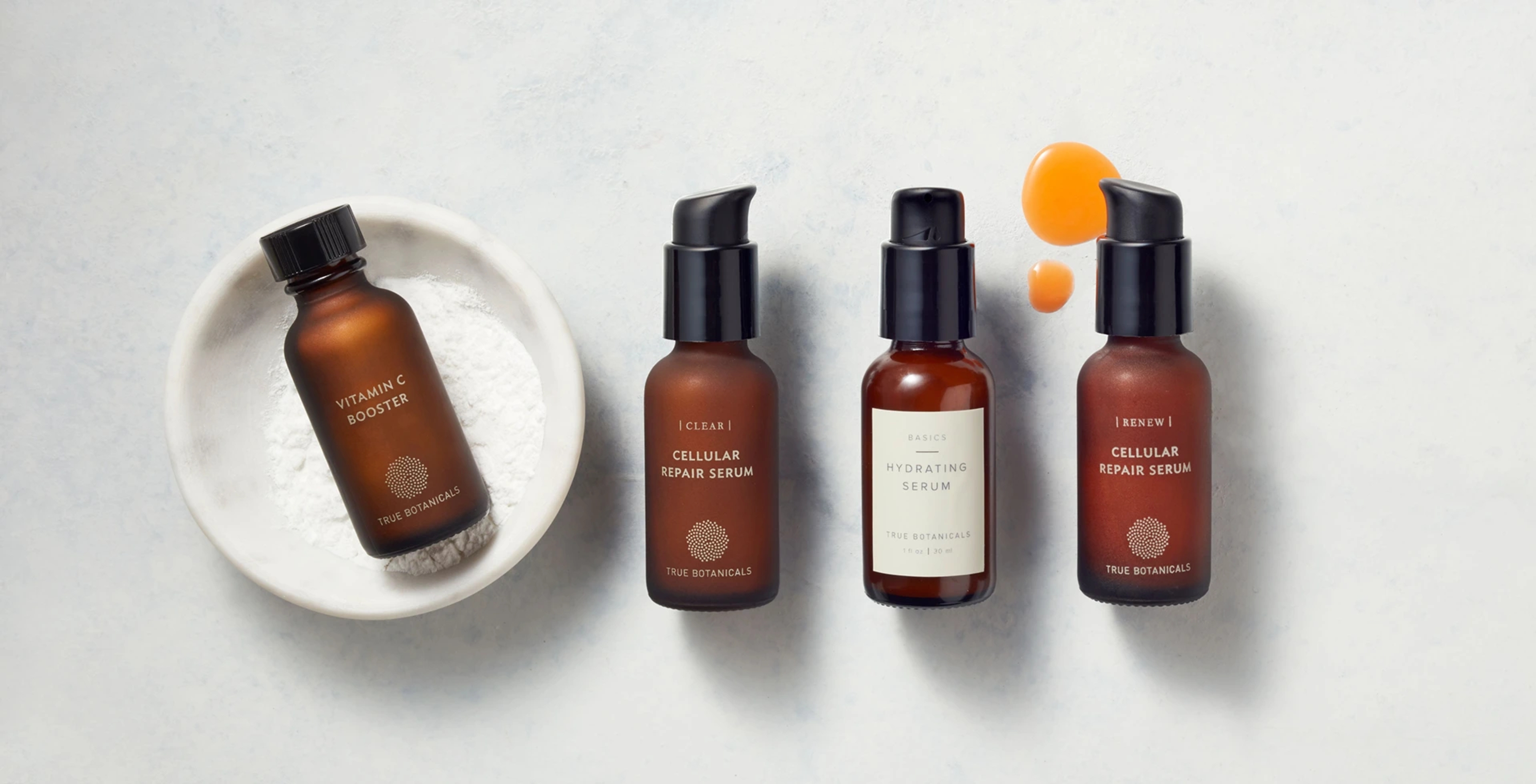 A set of True Botanicals products in brown bottles on a white table with a bit of orange product on the surface.