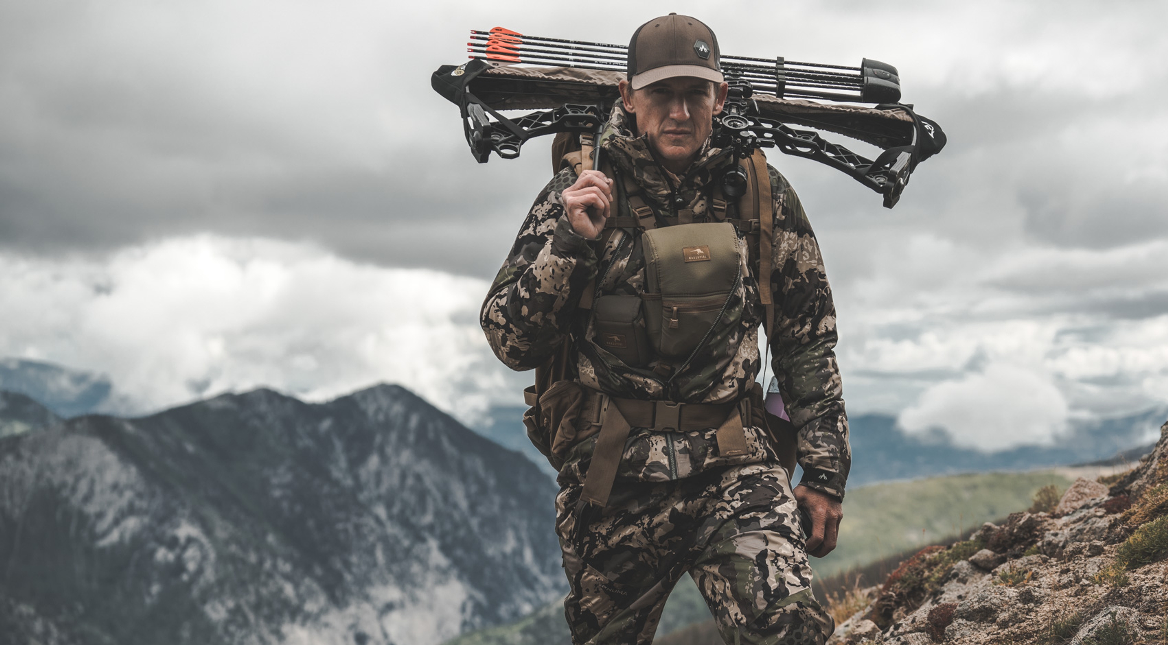 A man in camouflage holding hunting gear over his shoulder on a mountain
