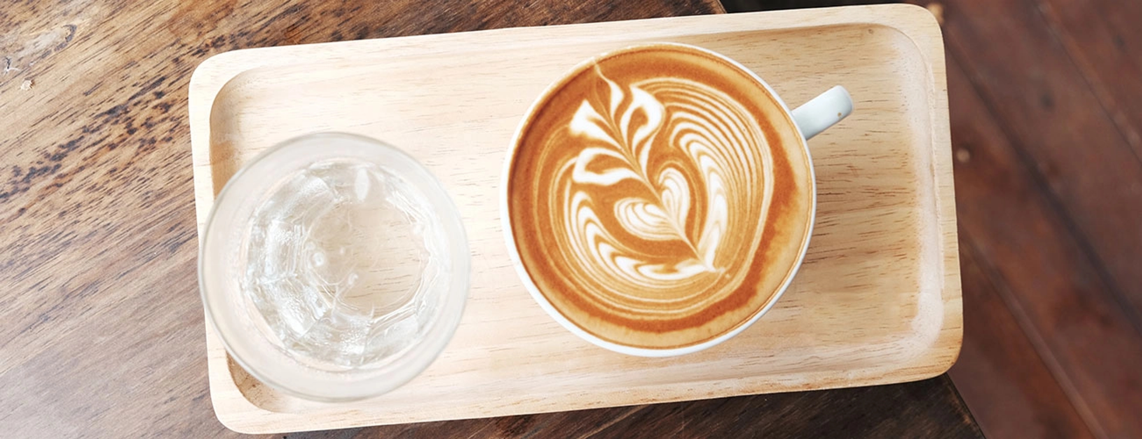 A shallow wooden serving plate with a glass of ice water and a coffee sitting on a wooden table