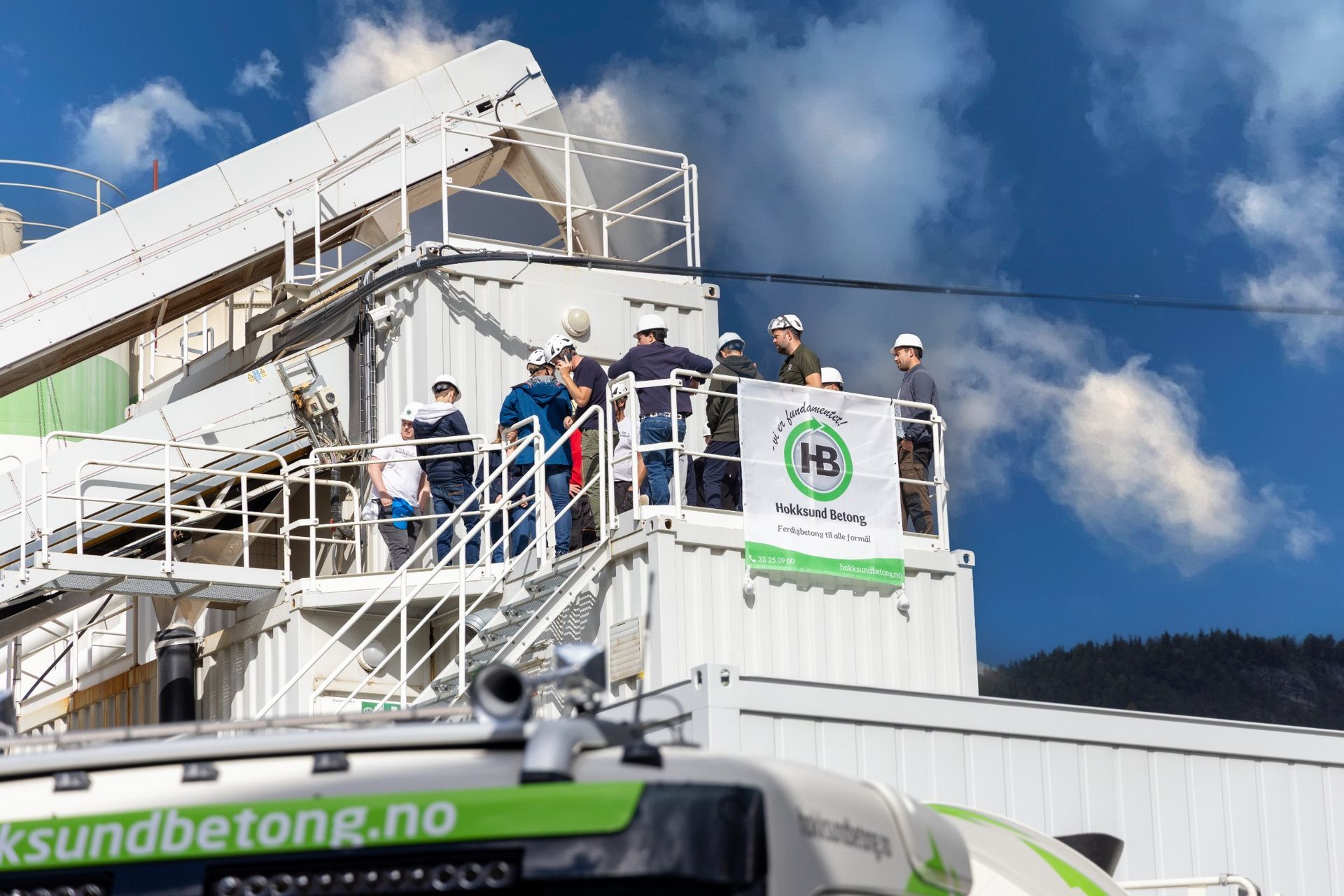 Ansatte med hvite hjelmer samlet på en plattform ved et betongverk under blå himmel, med banner for Hokksund Betong