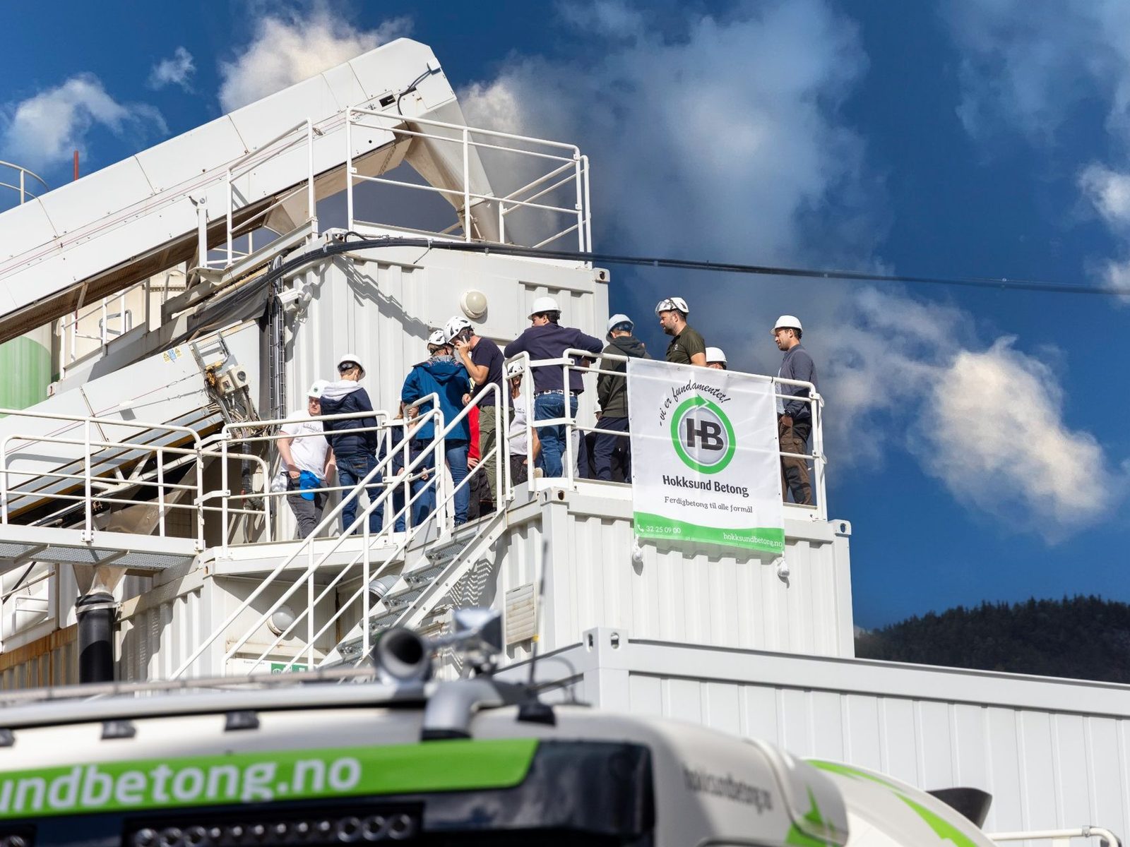 Ansatte med hvite hjelmer samlet på en plattform ved et betongverk under blå himmel, med banner for Hokksund Betong