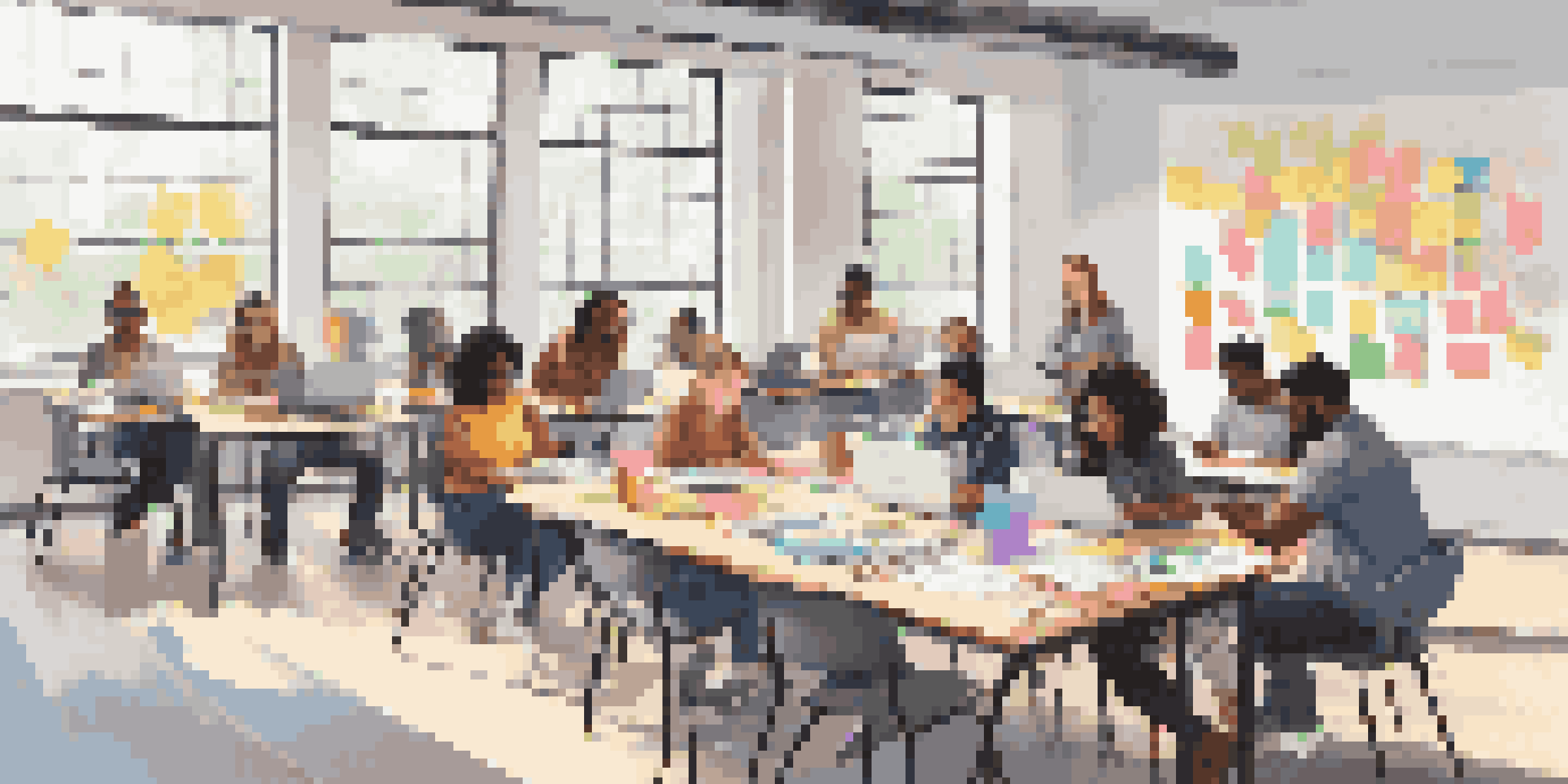A diverse group of students working together at a table, using laptops and digital tools in a bright classroom.