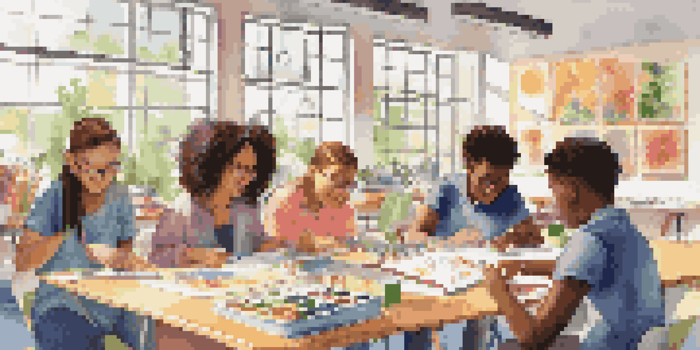 A diverse group of students working together at a table in a bright classroom filled with art supplies and laptops, with sunlight streaming in through windows.