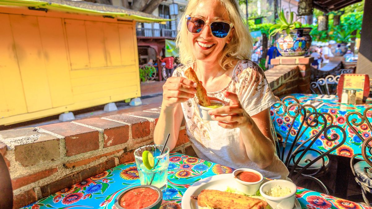 Joyful blonde woman puts enchilada in chili sauce, at typical Mexican food, at traditional restaurant of El Pueblo, Olvera street, Los Angeles Downtown State Historic Park, California, United States.