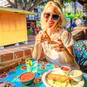 Joyful blonde woman puts enchilada in chili sauce, at typical Mexican food, at traditional restaurant of El Pueblo, Olvera street, Los Angeles Downtown State Historic Park, California, United States.