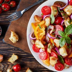 delicious fresh classic italian salad panzanella with tomatoes, croutons basil and onion rings on a white plate on an old wooden table with ingredients, view from above, flat lay, close-up