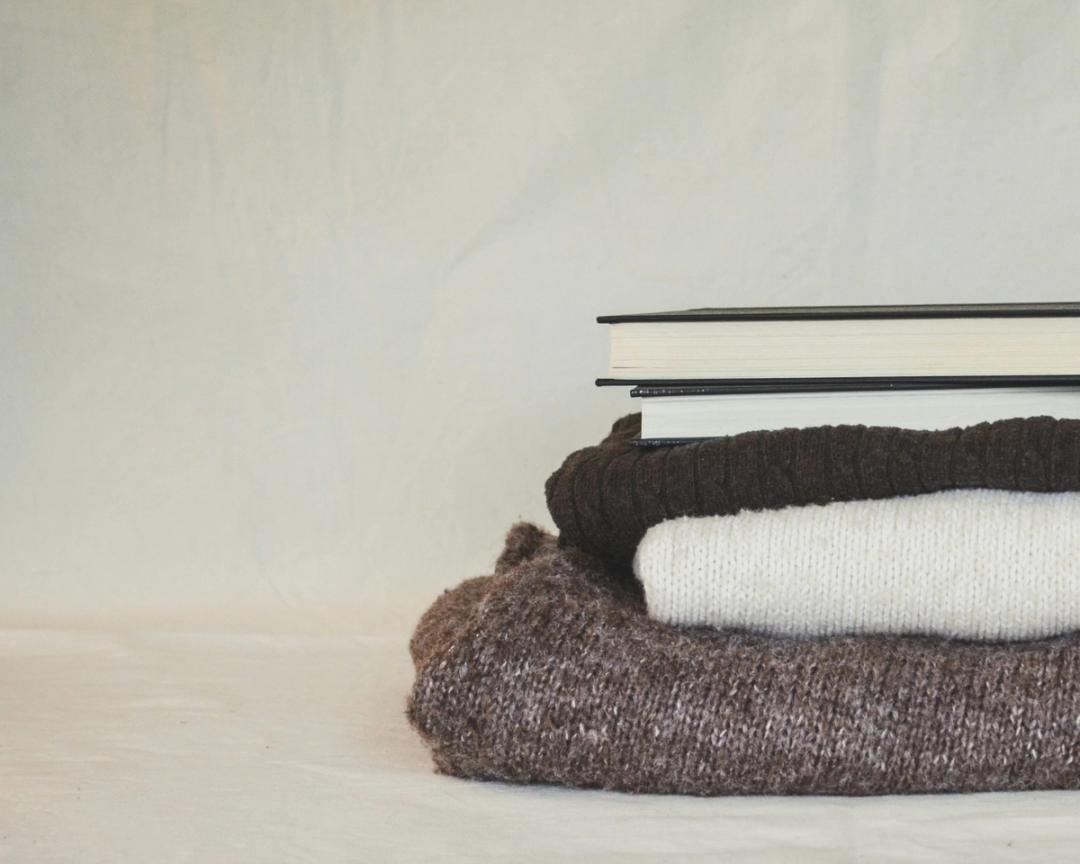 Zwei gebundene Bücher auf drei gefalteten Strickpullovern vor einem schlichten cremefarbenen Hintergrund.