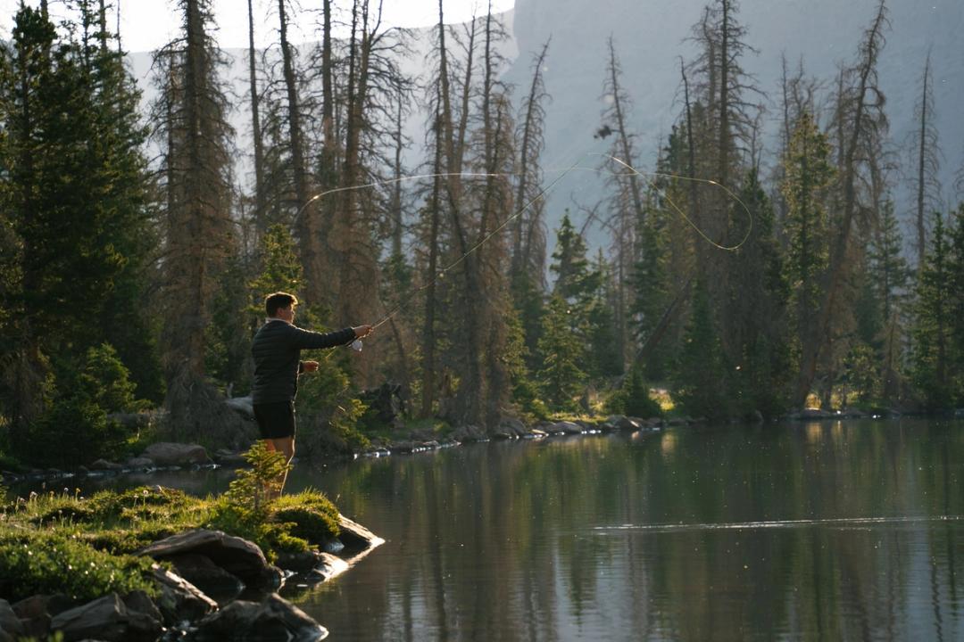 Un homme lance sa ligne de pêche à la mouche depuis une rive rocheuse, sur un lac de montagne calme bordé de pins.