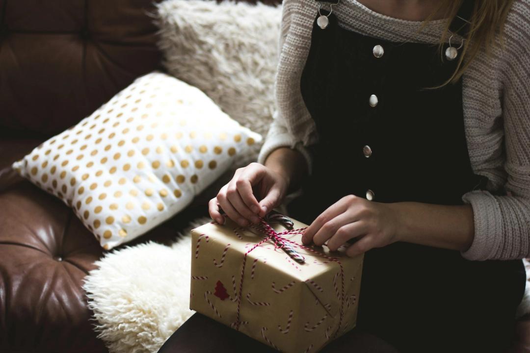 Des mains tiennent la ficelle d'un colis emballé dans du papier kraft, posé sur les genoux de quelqu'un, dans un canapé en cuir garni de coussins.