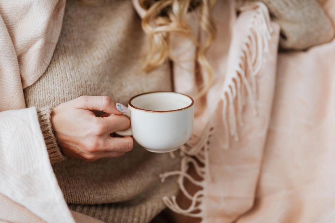 A person wrapped in a knitted jumper and a soft fringed blanket holds a small white cup.