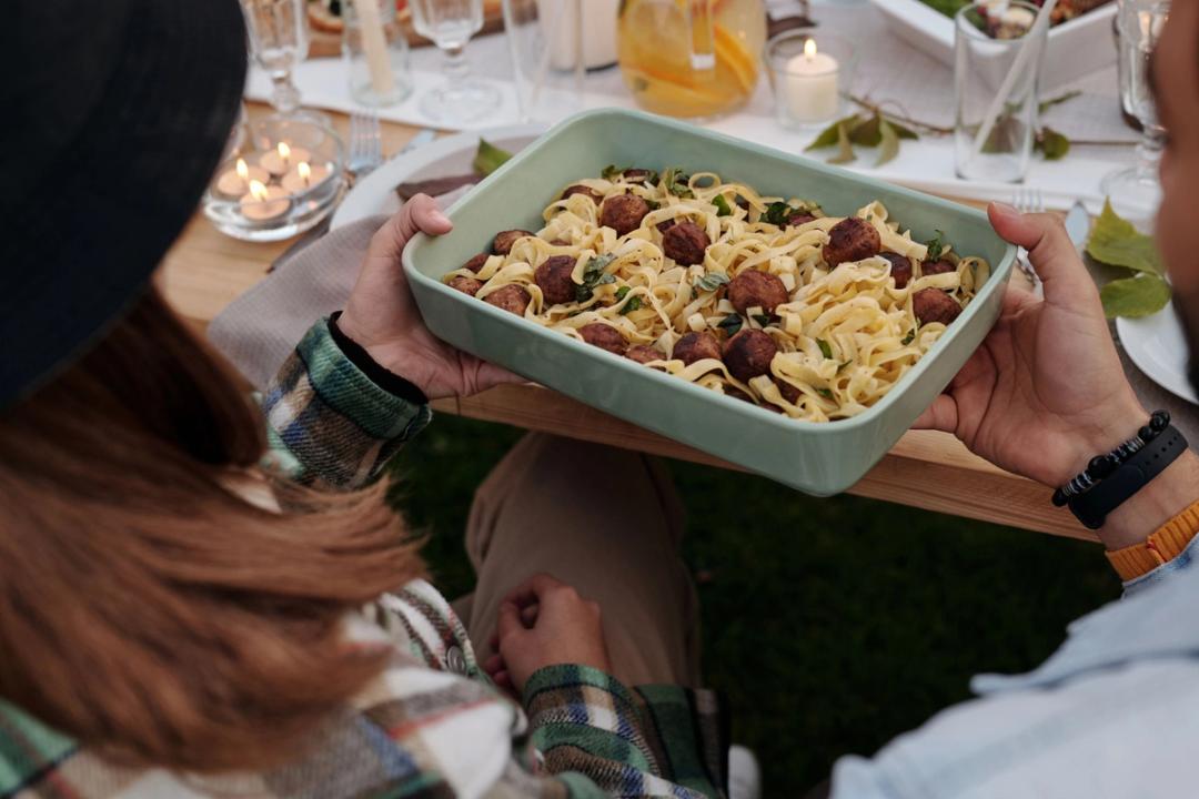 Des mains se passent un plat de tagliatelles aux boulettes de viande, autour d'une table dressée en plein air et éclairée à la bougie.
