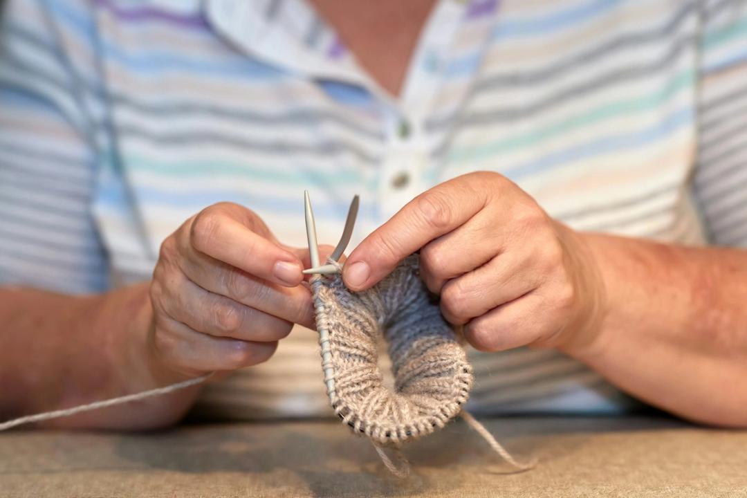 Hands knitting a grey sock on fine needles in front of a pastel striped shirt.