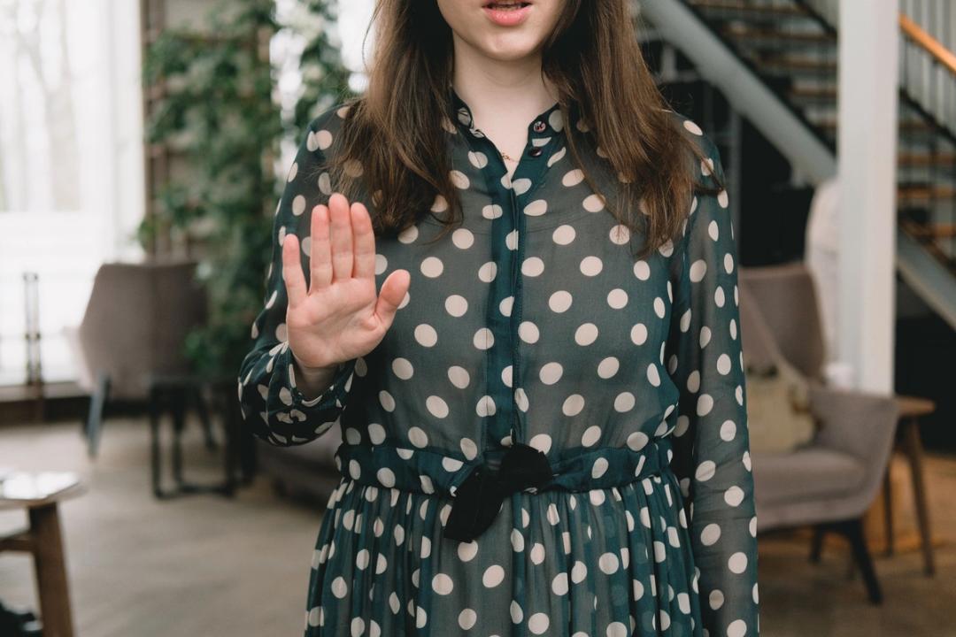A woman in a green polka-dot dress, her face cropped at the mouth, holds one hand up with the palm out in a bright room.