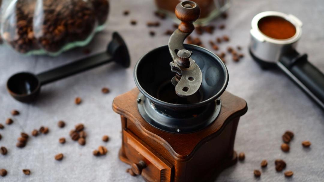 A wooden hand grinder with a crank handle among scattered coffee beans, a scoop and a filled portafilter beside it.