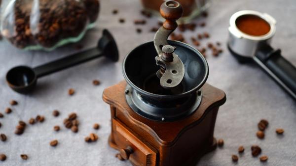 Eine hölzerne Handmühle mit Kurbel zwischen verstreuten Kaffeebohnen, daneben ein Messlöffel und ein gefüllter Siebträger.