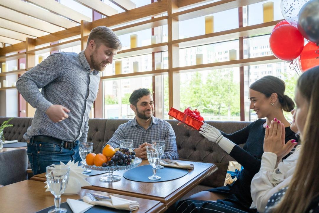 At a restaurant table by a window, a man presents a red gift box with a bow to a smiling woman, who reaches for it with a white prosthetic hand, while another woman claps.