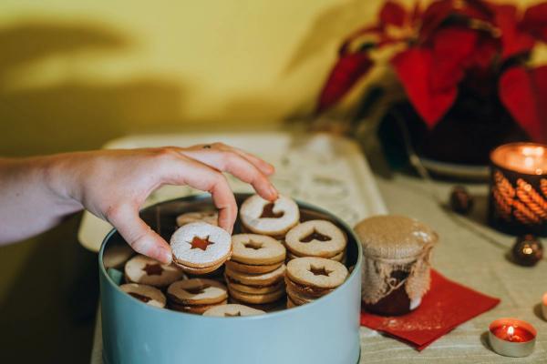 Une main soulève d'une boîte ronde un sablé en forme d'étoile fourré à la confiture, devant une bougie allumée et un poinsettia.