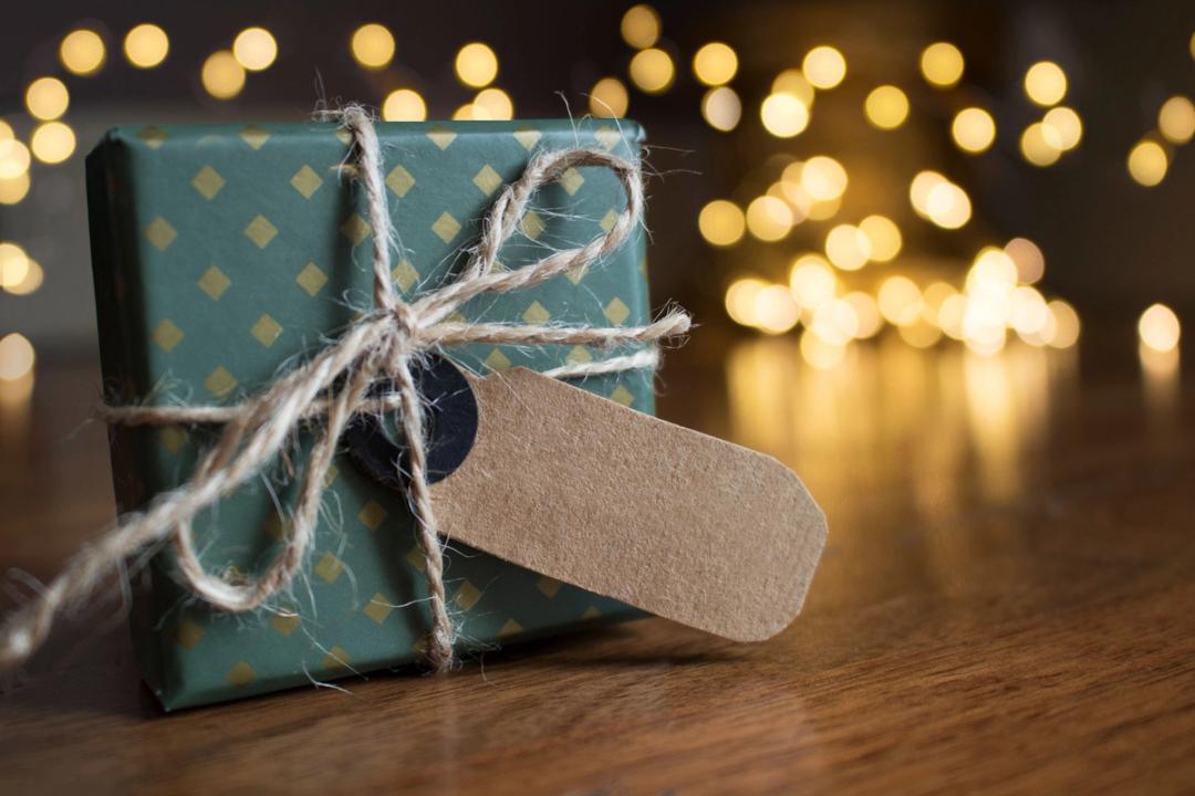 Un petit cadeau emballé dans du papier vert à losanges dorés, noué de ficelle avec une étiquette kraft vierge, sur une table en bois devant des lumières chaudes floues.