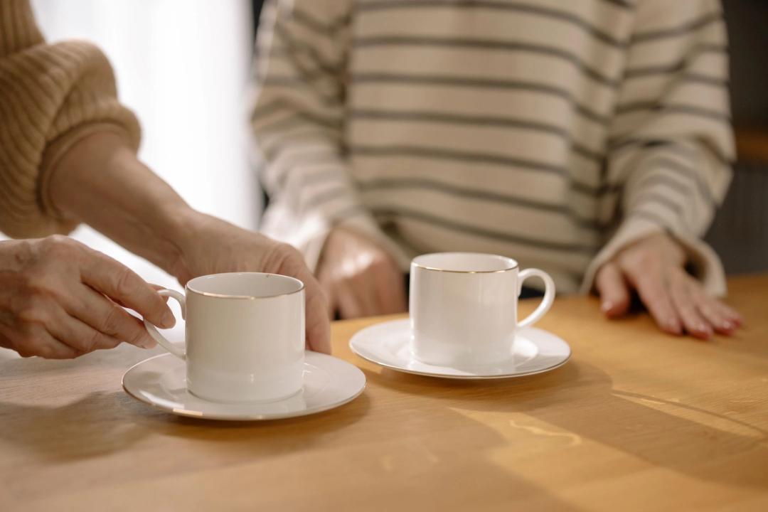 Two people in knitted jumpers at a wooden table, each with a white cup and saucer.