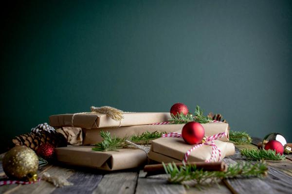 Pakete in Packpapier, mit Kordel und rot-weißem Garn verschnürt, dazu rote Christbaumkugeln, Tannenzweige und Zapfen auf einem rauen Holztisch vor einer dunkelgrünen Wand.