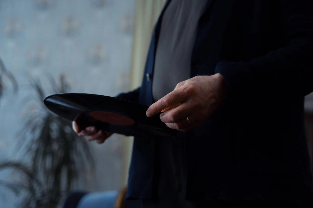 A man's hands holding a vinyl record in a dimly lit room, a houseplant behind him.